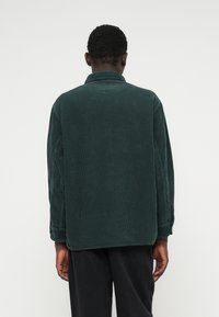 Camisa de corduroy verde escuro com mangas longas, punhos com botão e um corte descontraído. Apresenta uma textura ribbed vertical e um colarinho clássico.