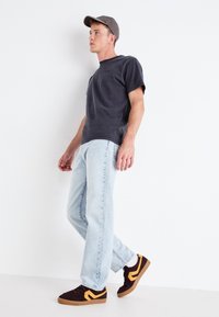 Dunkelgraues T-Shirt, hellblaue Jeans und braune Sneakers mit gelben Akzenten. Modell trägt eine passende Mütze und steht vor einem weißen Hintergrund.