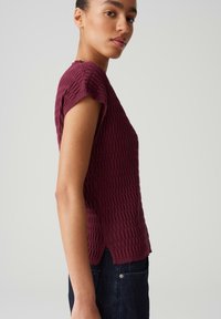 Burgundy textured short-sleeve top with a subtle ribbed pattern, side slits, and a relaxed fit. Paired with dark denim jeans.