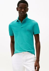 Mann in türkisfarbenem Poloshirt mit kleinem Logo und weißen Hosen, der mit Händen in den Taschen vor einem einfarbigen Hintergrund steht.