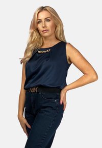 Granatowa satynowa bluzka bez rękawów z złotym łańcuchem na dekolcie, zestawiona z ciemnymi, wysokimi dżinsami i czarnym paskiem.