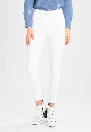 Femme portant un jean skinny blanc, une chemise en jean bleue avec des détails usés et des bottines blanches avec des œillets dorés.