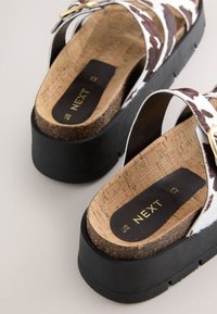 Korgist platvormsandaalid musta kummist tallaga, millel on laiad pruunide ja valgete lehmamustriga rihmad ning kuldse pandlaga aktsent.