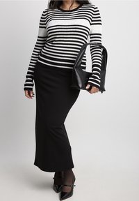 Gestreifter, schwarz-weißer Rippstrickpullover kombiniert mit einem schwarzen, taillierten Rock und glänzenden, schwarzen High Heels. Das Model hält eine schwarze Tasche.