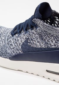 Námořnická pletená sportovní obuv s texturovaným svrškem, hladkým tmavě modrým akcentem a bílou mezipodešví s viditelnou vzduchovou jednotkou.