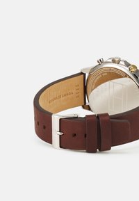 Cinturino in pelle marrone con fibbia in argento, caratterizzato da una cassa rotonda in acciaio inossidabile e un fondello in vetro trasparente. Dettagli in cucitura marrone visibili.