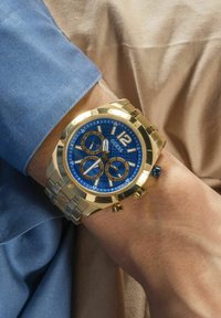 Auksinės spalvos chronograph laikrodis su mėlynu ciferblatu, auksiniais akcentais, keliais ciferblatais ir nerūdijančio plieno apyranke. Nešiojamas ant riešo, prilietus prie audinio.