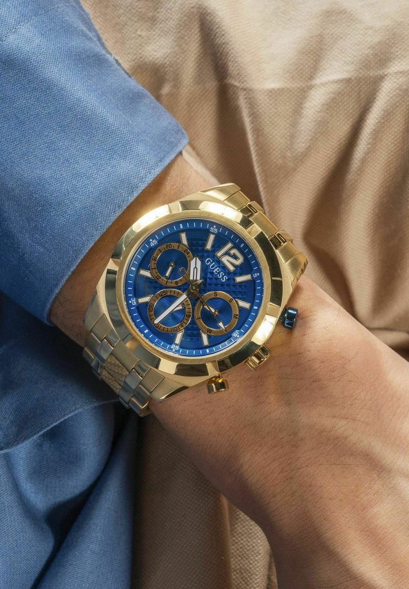Auksinės spalvos chronograph laikrodis su mėlynu ciferblatu, auksiniais akcentais, keliais ciferblatais ir nerūdijančio plieno apyranke. Nešiojamas ant riešo, prilietus prie audinio.