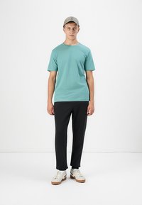 Tânăr îmbrăcat cu un tricou verde deschis, pantaloni negri, adidași albi și o șapcă bej, stând pe un fundal alb simplu.