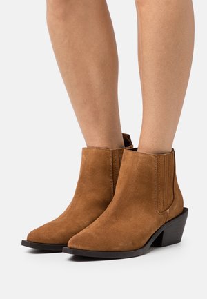 Bottines en suède marron avec un bout pointu, des panneaux latéraux élastiques et un petit talon empilé. Texture lisse avec des détails matériels minimaux.