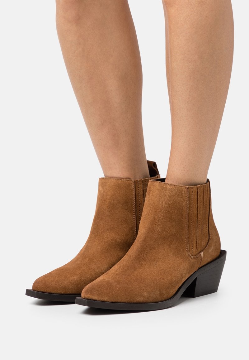 Bottines en suède marron avec un bout pointu, des panneaux latéraux élastiques et un petit talon empilé. Texture lisse avec des détails matériels minimaux.