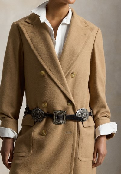 Nő, aki tevebarna kétsoros kabátot visel fehér inggel és fekete övvel, melyen nagy ezüst színű díszes csatok vannak.