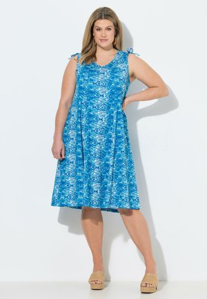 Kobieta ubrana w niebieską, wzorzystą sukienkę bez rękawów, sięgającą do kolan, z wiązanymi ramiączkami, stoi z jedną ręką na biodrze i beżowymi sandałami na koturnie.