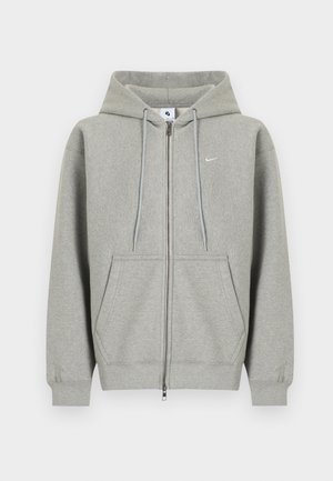 Sweat à capuche gris zippé en matière douce, avec une poche avant, une capuche à cordon et un petit logo blanc sur le côté gauche de la poitrine.