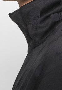 Svart jacka med hög krage, mjukt tyg och subtilt präglat logomönster. Rena linjer och ingen synlig hårdvara eller detaljer.