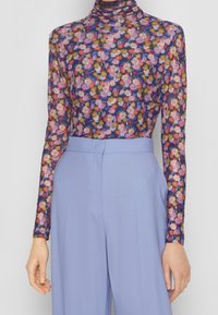 Długi rękaw bluzka z przezroczystego materiału w kwiatowy wzór z intensywnymi różowymi, fioletowymi i żółtymi kwiatami, zestawiona z wysokim stanem jasnoniebieskimi spodniami.