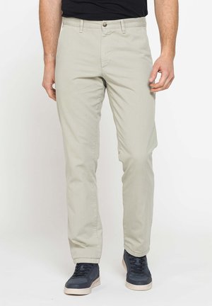 Uomo che indossa pantaloni beige chiaro dritti e sneakers nere, in piedi davanti a uno sfondo bianco uniforme.