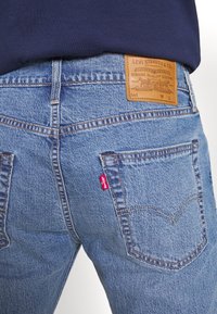 Vue arrière d'une personne portant un jean Levi's bleu avec un patch en cuir visible de la marque et une étiquette rouge avec logo sur la poche arrière droite.