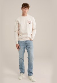 Šviesiai smėlinis džemperis su apvalia iškirpte ir tamsiai raudonu "Blue Ridge" grafiniu dizainu ant krūtinės, derinamas su šviesiai mėlynomis džinsomis ir baltais sportbačiais.