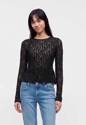 Ung kvinde med langt mørkt hår iført en sort blondebluse med lange ærmer og lyseblå jeans, stående mod en ensfarvet baggrund.