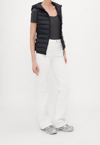 Svart vadderad väst med huva, bärs över en mörkgrå topp, kombinerad med vita byxor och grå sneakers, med en figurnära silhuett.