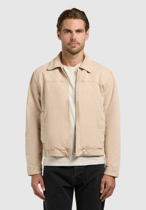 Man draagt een beige jas met ritssluiting over een wit shirt en zwarte broek, staand tegen een effen lichte achtergrond.