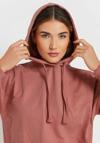 Felpa rosa polveroso con cappuccio regolabile, maniche lunghe e una texture morbida. Il modello sta tirando giù il cappuccio e guarda in avanti.