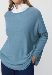 Jasnoniebieski, oversize sweter wykonany z miękkiego materiału, z szerokim dekoltem, prążkowanymi mankietami i luźnym krojem, noszony na białej koszuli z kołnierzykiem.