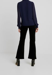 Blouse marine à manches bouffantes, associée à un pantalon évasé en velours noir et des bottes à talons noires. Textures lisses et coupe ajustée.