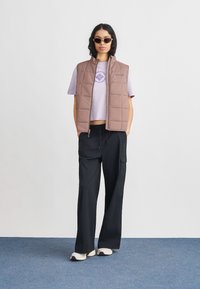 Γυναίκα που φοράει γυαλιά ηλίου, μπλούζα μανίκι κοντό σε μωβ απόχρωση, καφέ μπουφάν με καπιτονέ σχέδιο, μαύρο παντελόνι φαρδύ και λευκά αθλητικά, στέκεται σε μπλε χαλί.