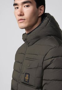 Giovane uomo che indossa una giacca trapuntata verde scuro con cappuccio e una tasca sul petto decorata con una piccola toppa a forma di cane.