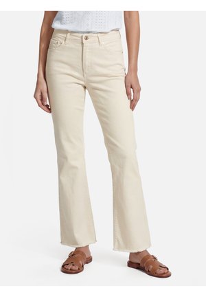Femme portant un jean large couleur crème avec des ourlets bruts et des sandales plates en cuir marron sur fond blanc.