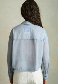 Camicia corta azzurra con colletto, polsini abbottonati e dettaglio a toppa sul retro. Realizzata in un tessuto leggero con una texture liscia.