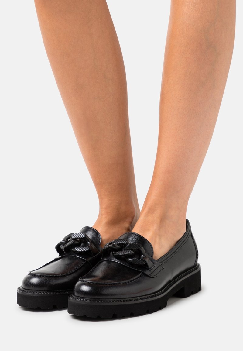 Paire de mocassins en cuir noir avec semelles épaisses et grand détail chaîne décoratif, portés sur jambes nues.