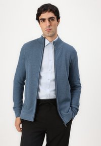Blue zip-up cardigan in textured knit, featuring a stand collar, long sleeves, and side pockets, worn over a light blue shirt.