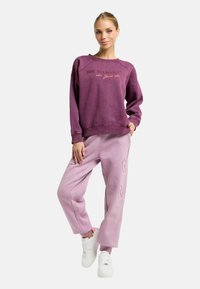 Paarse trui met ronde hals en reliëftekst; gecombineerd met lichtroze joggers met een zijlogo. Witte sneakers maken de look compleet.