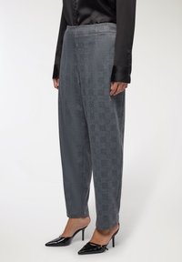 Osoba ubrana w szare w kratę spodnie, czarną satynową bluzkę i czarne szpilki na wysokim obcasie stoi na białym tle.