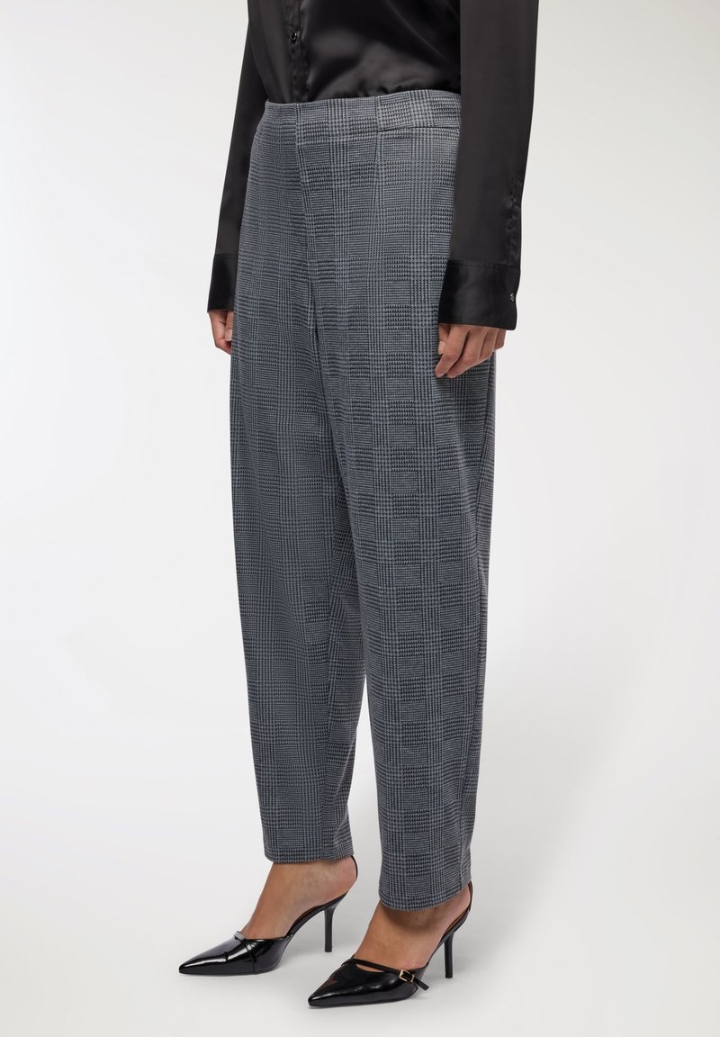 Osoba ubrana w szare w kratę spodnie, czarną satynową bluzkę i czarne szpilki na wysokim obcasie stoi na białym tle.