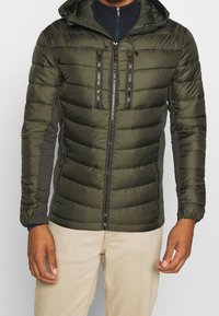 Veste rembourrée de couleur vert foncé avec fermeture éclair à l'avant. Comprend deux poches poitrine, des poignets élastiques et une capuche. Panneaux latéraux en tissu gris.