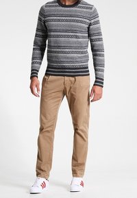 Pull gris foncé à motifs avec des dessins géométriques gris clair et blancs, assorti à un pantalon chino beige et des chaussures blanches avec des rayures rouges.