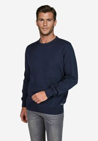 Marineblauwe trui met een ronde hals, lange mouwen, geribbelde manchetten en zoom, gemaakt van een zachte stof. Gecombineerd met lichtgrijze jeans.