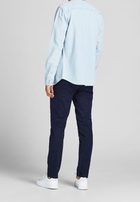 Homme portant une chemise bleu clair à manches longues, un pantalon bleu foncé et des baskets blanches, debout de dos sur un fond clair uni.