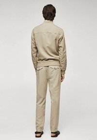 Tuta in cotone beige con giacca aderente e pantaloni tapered, dotato di tasche posteriori e una sottile texture. Indossato con sandali senza strap.