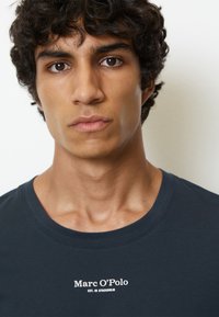 Dunkelblaues T-Shirt mit Rundhalsausschnitt; verfügt über das weiße Textlogo "Marc O’Polo EST. IN STOCKHOLM", das mittig auf der Brust platziert ist.