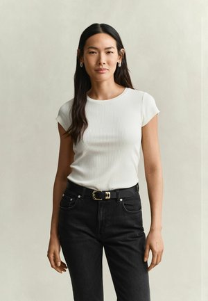 Jeune femme aux longs cheveux foncés portant un haut blanc à manches courtes côtelé et un jean noir avec une ceinture noire, debout devant un fond uni.