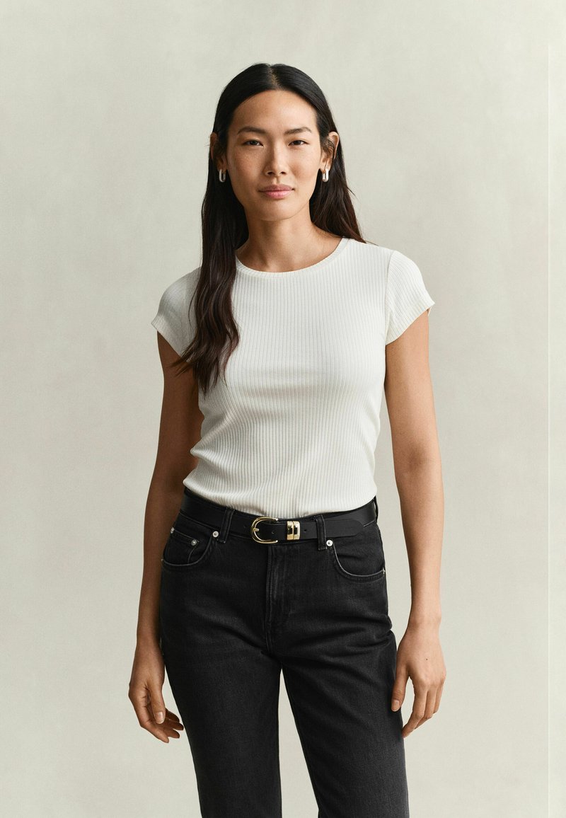 Jeune femme aux longs cheveux foncés portant un haut blanc à manches courtes côtelé et un jean noir avec une ceinture noire, debout devant un fond uni.