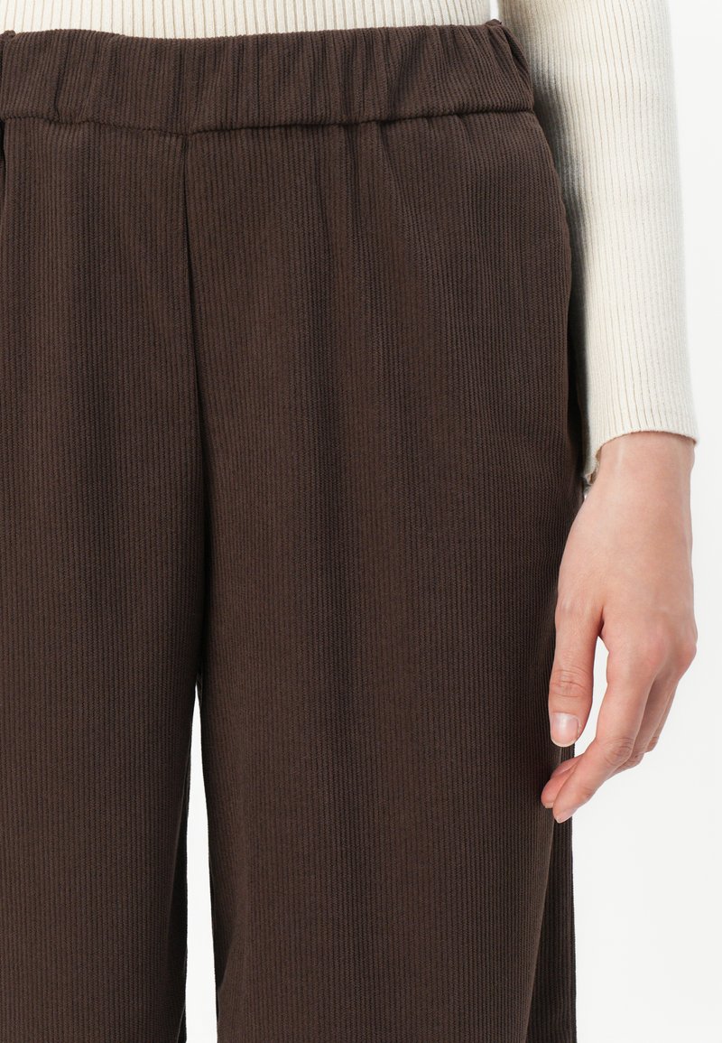 Close-up van een persoon in ribbelige donkerbruine broek met elastische tailleband en een crëmekleurig ribbelig lange mouwen tops.