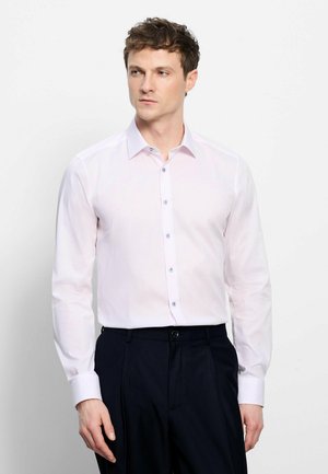 Uomo che indossa una camicia rosa chiaro a maniche lunghe con bottoni e pantaloni scuri, in piedi davanti a uno sfondo bianco uniforme.