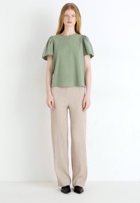 Svetlozeleno kratkotrajno majico; sproščen kroj z naborki na rokavih. V kombinaciji s svetlo bež širokima hlačami. Črne čevlje z ostrim nožnim delom. Enostavna ozadje.