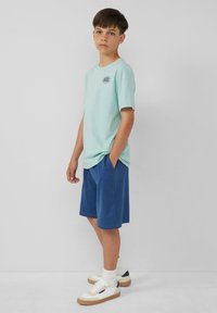 Lichtblauw katoenen T-shirt met een klein zwart logo, gecombineerd met donkerblauwe shorts en witte sneakers met tan zolen en marineblauwe accenten.
